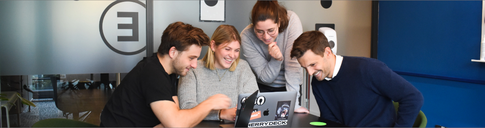 Four people sit in an office environment around a laptop. They are similing.